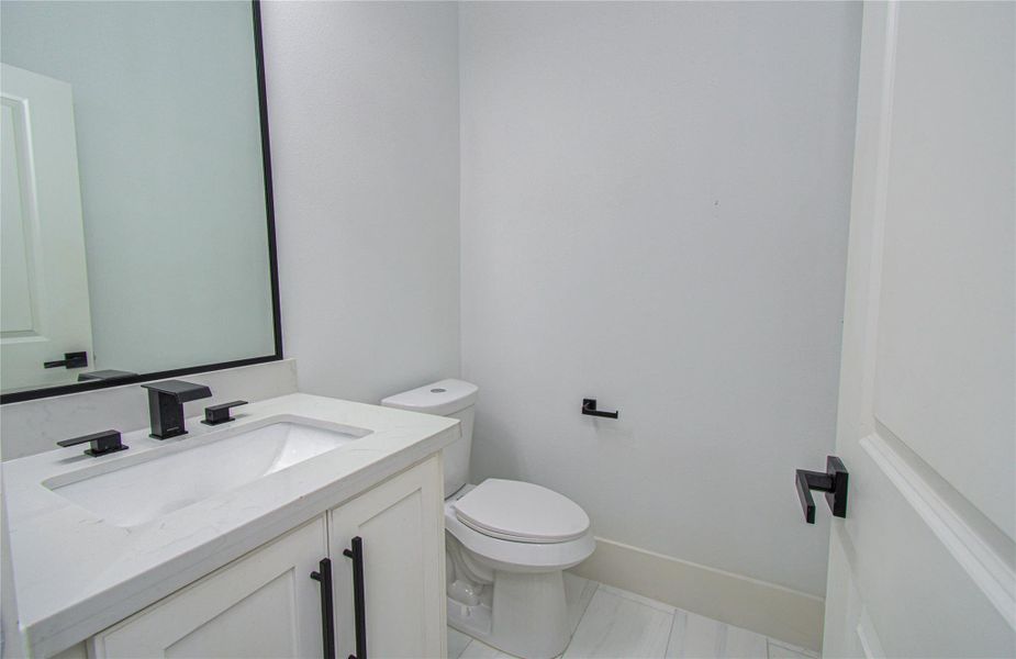 The modern half-bathroom located downstairs for easy entertaining features a sleek white vanity with black fixtures, a large mirror, and a toilet. The room is bright and has a clean, contemporary design.