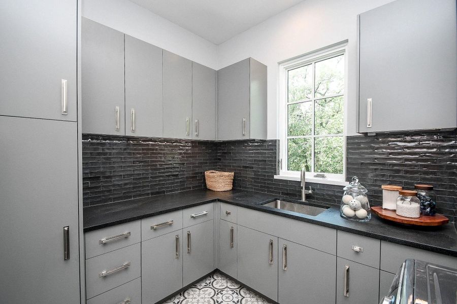 This modern kitchen features sleek gray cabinetry, black countertops, and a stylish tile backsplash. It has ample storage, a large window for natural light, and elegant patterned floor tiles, creating a functional and chic space.