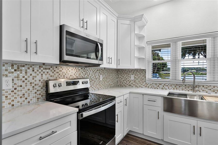 The space features pristine white cabinetry that provides ample storage, paired with elegant marble-like countertops that offer plenty of room for meal preparation. The mosaic tile backsplash adds a touch of sophistication and color contrast, enhancing the overall aesthetic.Equipped with modern appliances, the kitchen includes a sleek black cooktop and a matching microwave, seamlessly integrated into the cabinetry for a streamlined look. A refrigerator is also included, making this kitchen ready for immediate use upon purchasing the home. A large window over the sink allows for natural light to flood the space, creating a warm and inviting atmosphere. This kitchen combines practicality with elegant design, making it a standout feature of the home.