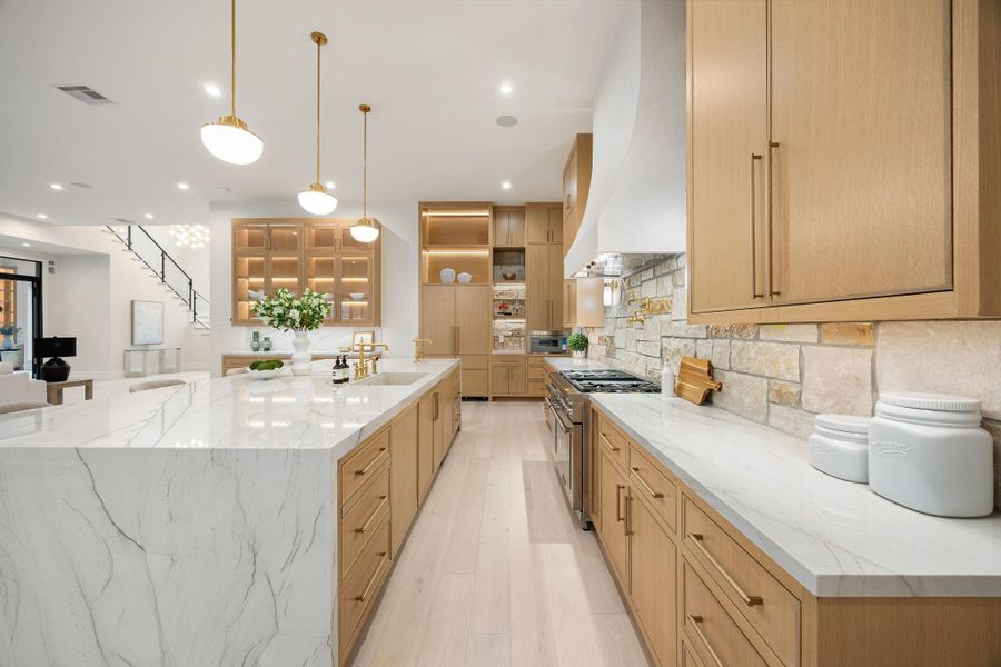A refined galley-style perspective showcases the length and functionality of this beautifully designed kitchen. Clean countertops, custom cabinetry, and warm finishes create a cohesive look throughout. The highlight is the extended island workspace, offering both prep and seating, while maintaining an open sightline to the surrounding living areas, enhancing flow and connectivity.