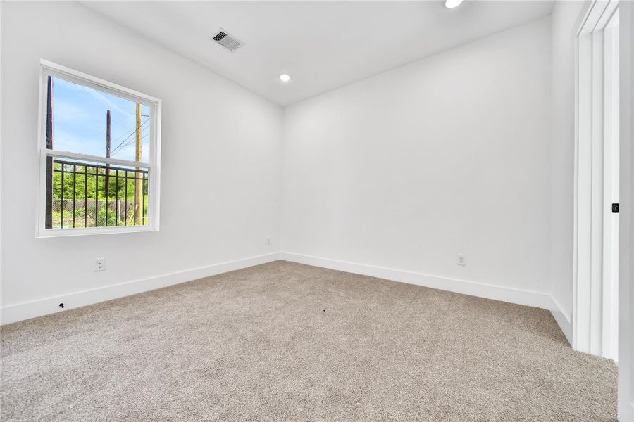 This room features clean white walls, recessed lighting, and a carpeted floor. A window allows natural light in, offering a view of greenery outside. Perfect for a cozy bedroom or home office.