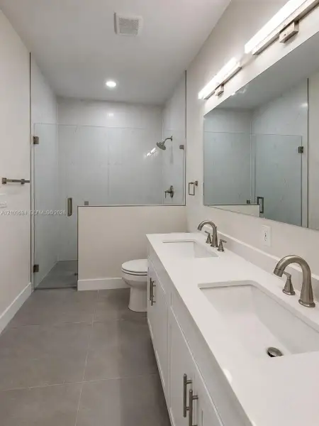Double vanity with white quartz countertops and undermount sinks, wall-to-wall vanity mirror, large-format tile flooring, and walk-in shower with glass enclosure
