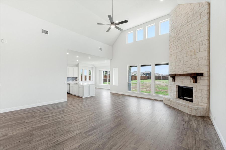 Open living room with vaulted ceiling, stone fireplace, hardwood floors, and open kitchen in background