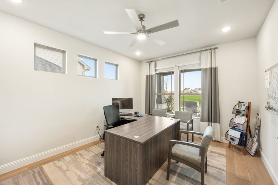 Home office with L-shaped desk, ceiling fan, hardwood floors, and large windows with gray curtains