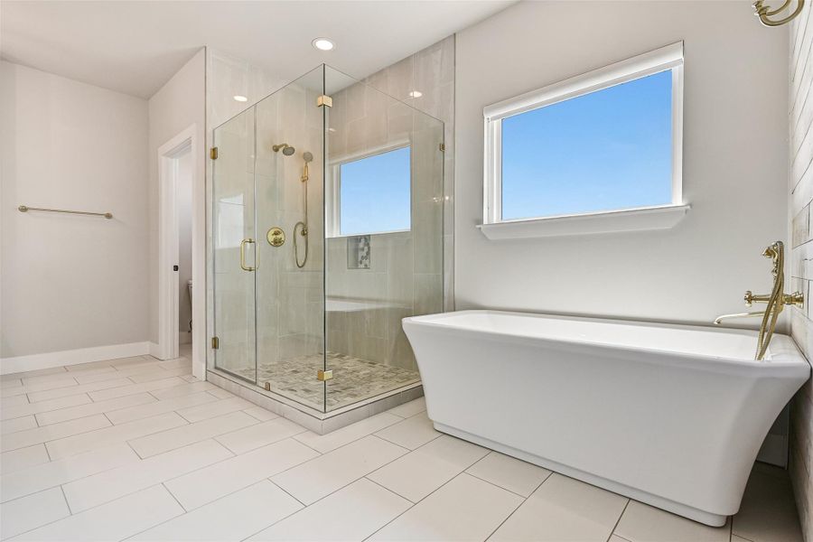 A freestanding soaking tub beneath oversized windows creates a calming focal point within the primary bath. Paired with a spacious glass shower and elegant finishes, the space feels refined, airy, and indulgent.