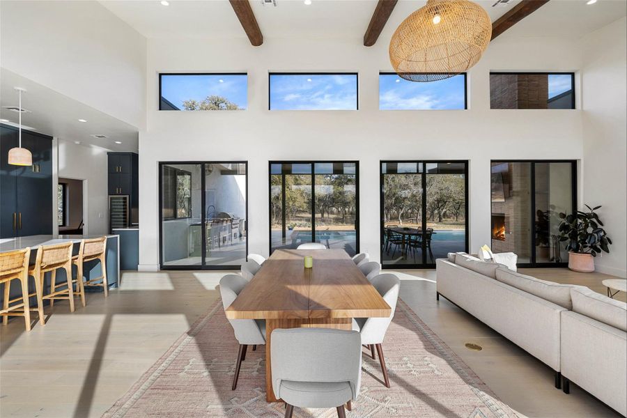 Dining room featuring beam ceiling, light wood-style floors, recessed lighting, a towering ceiling, and a warm lit fireplace
