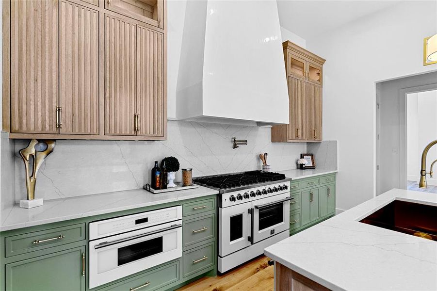 Kitchen with light stone counters, wall chimney exhaust hood, gas range, green cabinets, and decorative backsplash Kitchen with light stone counters, wall chimney exhaust hood, gas range, green cabinets, and decorative backsplash