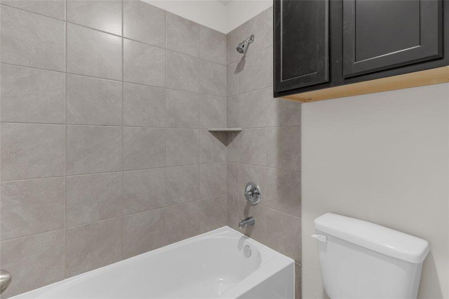 This is a modern bathroom with large, grey tiles, featuring a combined bathtub and shower area with a built-in niche for toiletries. Above the toilet, there is a dark cabinet for additional storage.