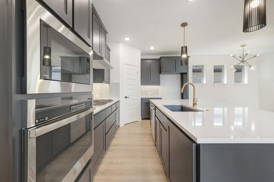 Contemporary kitchen featuring dark cabinetry, stainless steel appliances, a gas cooktop, white countertops, and a central island with an undermount sink and brass-finish faucet