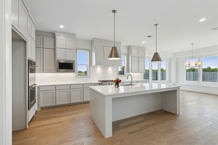 Open-concept kitchen featuring an oversized island with an integrated sink, light wood-finish flooring, recessed lighting, double wall ovens, and shaker-style cabinetry Open-concept kitchen featuring an oversized island with an integrated sink, light wood-finish flooring, recessed lighting, double wall ovens, and shaker-style cabinetry