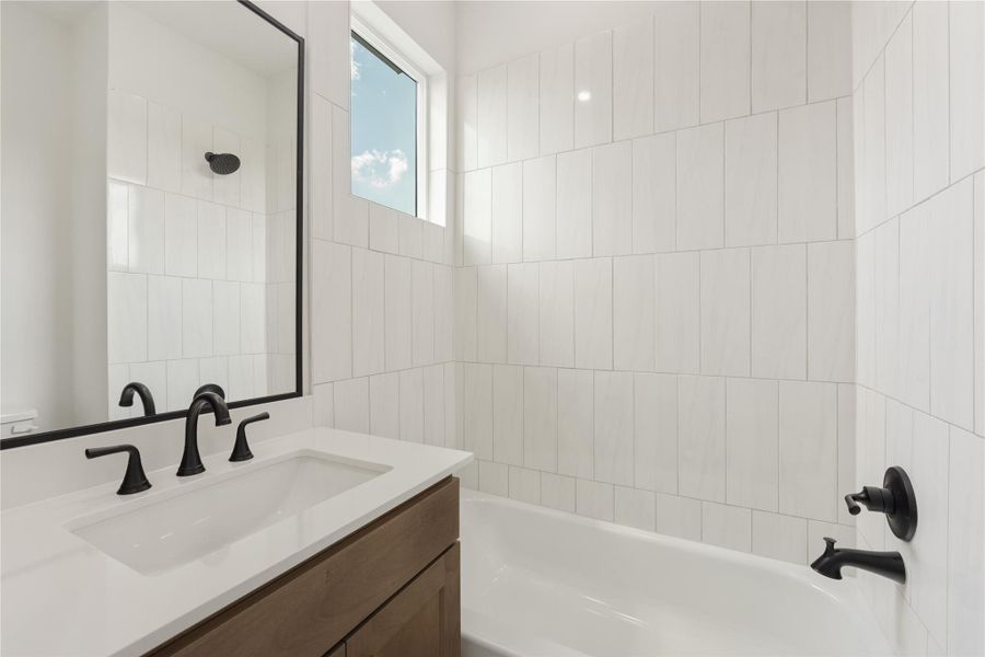 Secondary bath with a fresh modern design featuring a wood vanity, white countertop, black fixtures, framed mirror, tub/shower combination, and vertical tile surround. A high window brings in natural light, while the clean finishes and crisp contrast create a polished, functional bath.