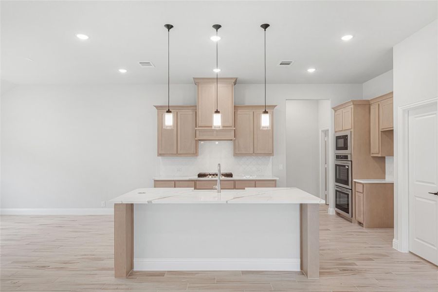 Kitchen with large white quartz island, light wood cabinets, three pendant lights, and light wood floors Kitchen with large white quartz island, light wood cabinets, three pendant lights, and light wood floors