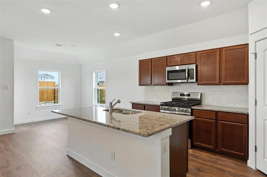 Kitchen with appliances with stainless steel finishes, backsplash, light stone countertops, dark wood finished floors, and a kitchen island with sink Kitchen with appliances with stainless steel finishes, backsplash, light stone countertops, dark wood finished floors, and a kitchen island with sink