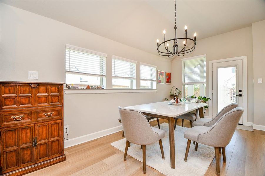Bright dining area with modern chandelier, large windows, and a door leading outside. Features a stylish table with four chairs and a classic wooden cabinet, creating a cozy yet elegant atmosphere.