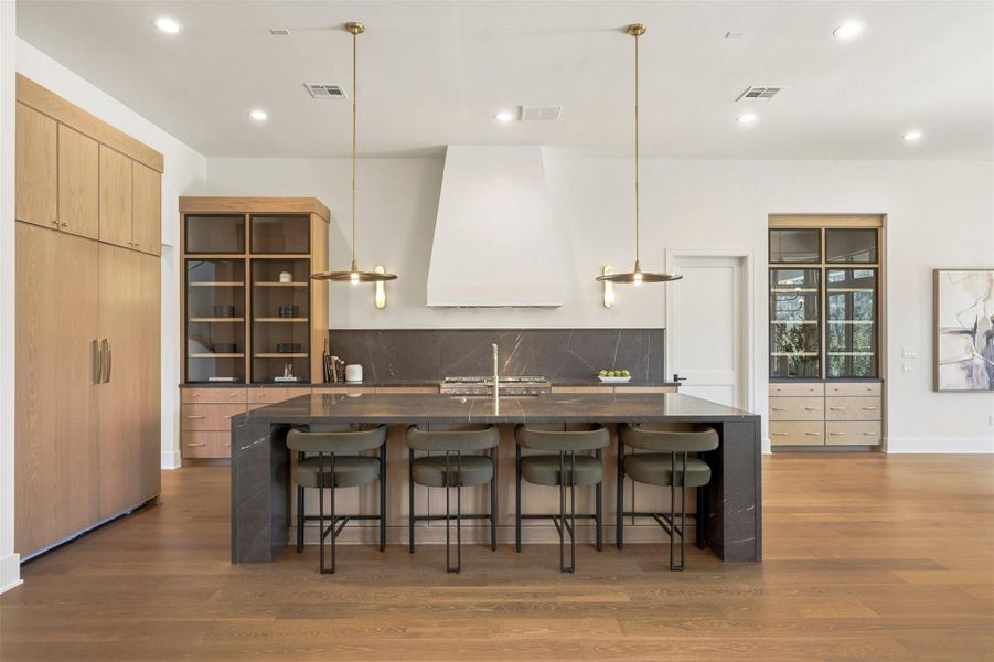 This modern kitchen features a sleek design with a large island, designer pendant lighting, and ample storage. Featuring a built-in stove, minimalistic shelving, and custom-paneled fridge. Closed door to the right leads to your chef's prep kitchen. This modern kitchen features a sleek design with a large island, designer pendant lighting, and ample storage. Featuring a built-in stove, minimalistic shelving, and custom-paneled fridge. Closed door to the right leads to your chef's prep kitchen.