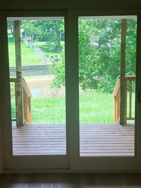 Sliding glass doors opening to a wood-finish deck with natural wood railings, overlooking a grassy expanse with mature trees and a body of water beyond