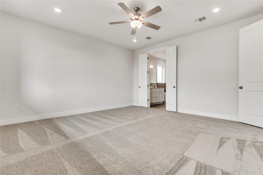 A spacious bedroom with plush carpet and direct access to a private bathroom, illuminated by recessed lighting and a ceiling fan.