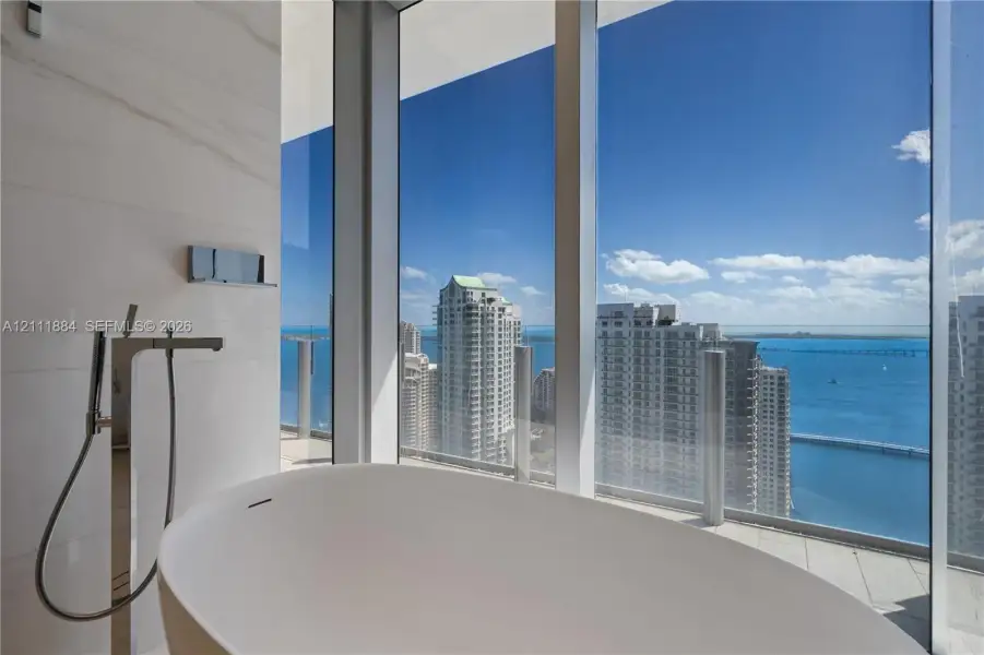 Floor-to-ceiling windows featuring panoramic water and city scenery, a white freestanding soaking tub, and wall-mounted metallic fixtures with a handheld shower wand Floor-to-ceiling windows featuring panoramic water and city scenery, a white freestanding soaking tub, and wall-mounted metallic fixtures with a handheld shower wand