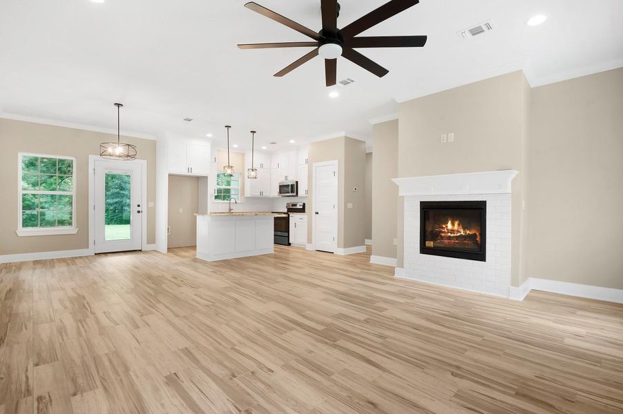 This photo showcases a spacious open-concept living area with light wood flooring and neutral walls. The room features a modern ceiling fan, a cozy fireplace, and ample natural light from large windows. The kitchen is equipped with stainless steel appliances, white cabinetry, and a central island with pendant lighting. A door leads to an outdoor space, enhancing the home's airy feel. This photo showcases a spacious open-concept living area with light wood flooring and neutral walls. The room features a modern ceiling fan, a cozy fireplace, and ample natural light from large windows. The kitchen is equipped with stainless steel appliances, white cabinetry, and a central island with pendant lighting. A door leads to an outdoor space, enhancing the home's airy feel.