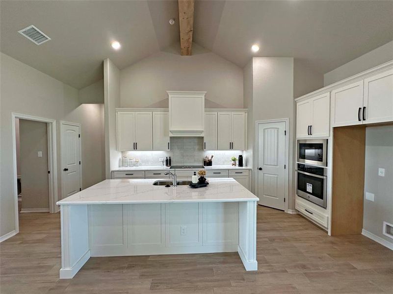 Kitchen featuring backsplash, white cabinetry, beamed ceiling, stainless steel oven, and a center island with sink Kitchen featuring backsplash, white cabinetry, beamed ceiling, stainless steel oven, and a center island with sink