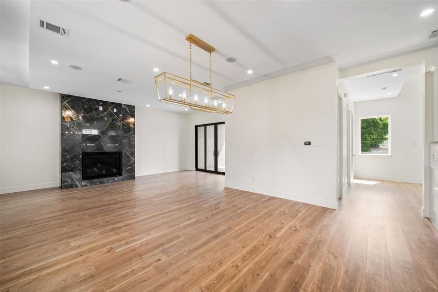 Spacious living area features a modern design with wood floors, a sleek black marble fireplace, and ample natural light. A chic gold light fixture adds elegance, while glass doors open to additional rooms, enhancing the open-concept layout.
