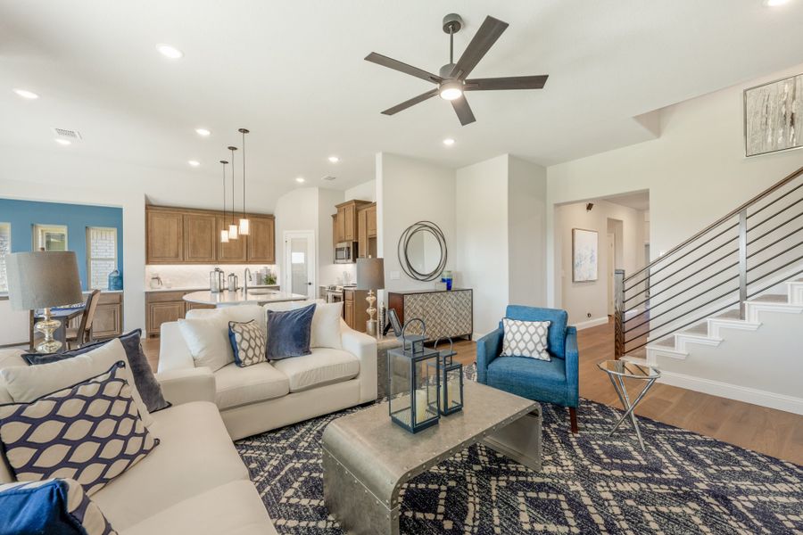 Open-concept living room with white sofas, blue accent chair, ceiling fan, and kitchen visible in background