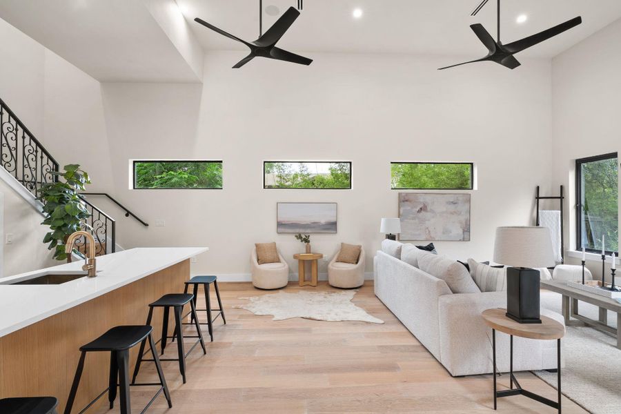 Open-concept living area featuring high ceilings with modern ceiling fans, wood-finish flooring, a kitchen island with seating, and a staircase with an ornate iron railing