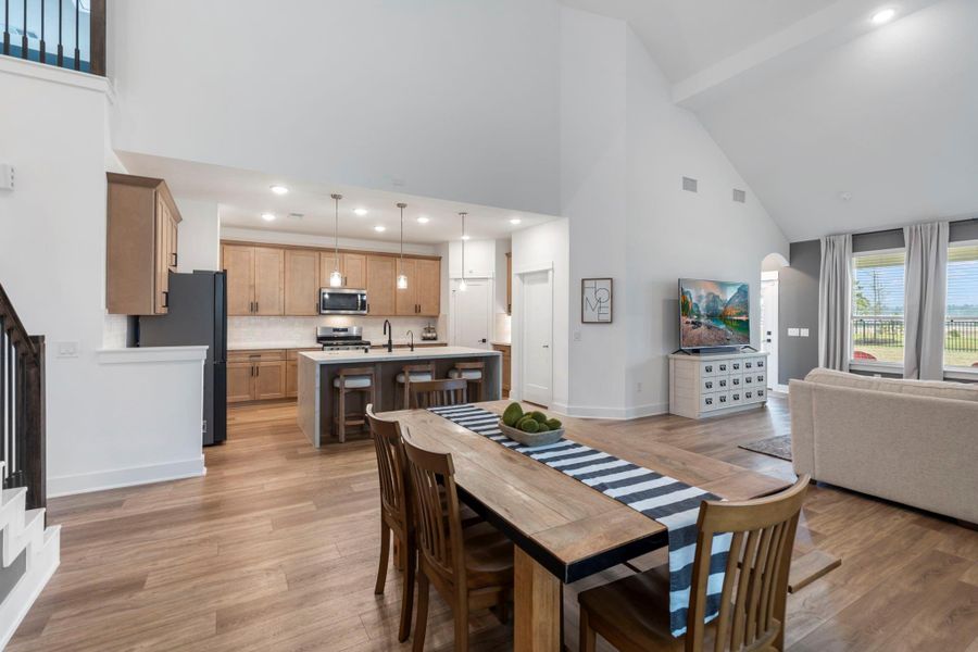 Spacious, open-concept living area features a modern kitchen with wood cabinets, a large island with bar seating, and high ceilings. The dining area and living room are bright and inviting, with large windows providing plenty of natural light and views of the outdoors.