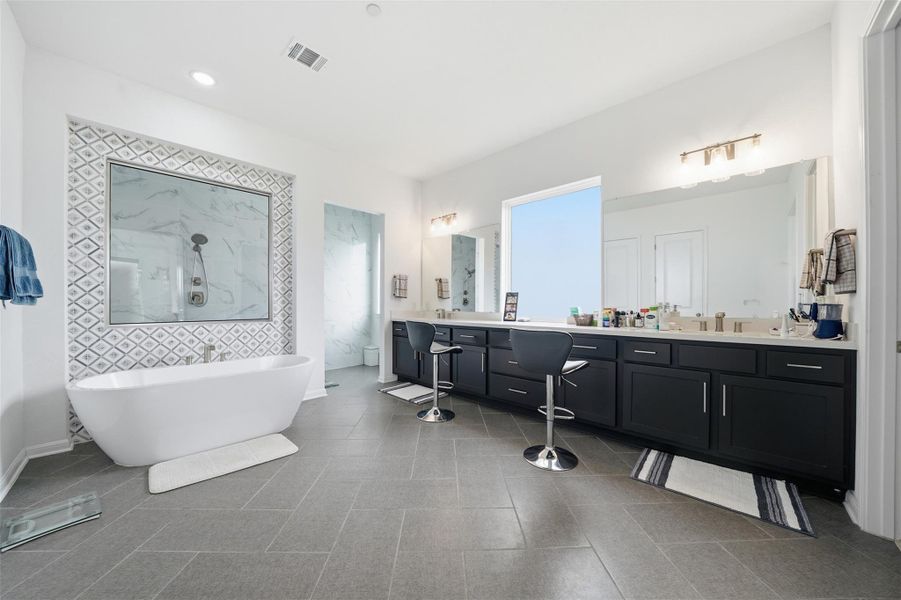 The bathroom features a freestanding tub, a large shower with a glass enclosure and decorative tile surround, a spacious vanity with dual sinks, and light gray tiled flooring