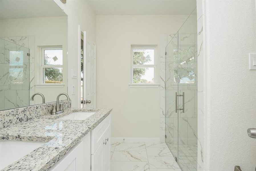 Modern bathroom with marble tiles, a glass shower door, granite countertop, and two windows allowing for natural light.