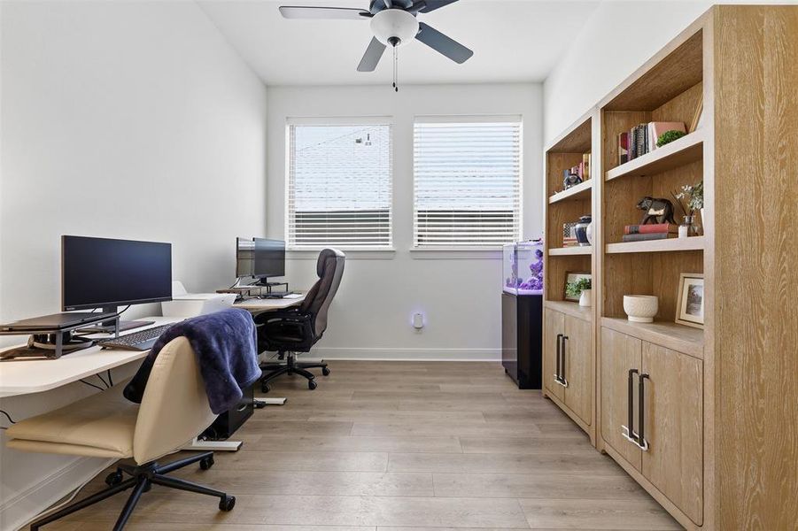 Bright room with dual windows, wood-finish flooring, a ceiling fan, and built-in shelving with lower cabinetry Bright room with dual windows, wood-finish flooring, a ceiling fan, and built-in shelving with lower cabinetry