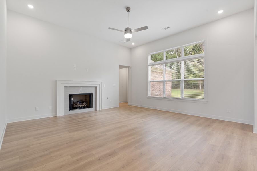 Spacious living area featuring a fireplace with a white mantel, wood-finish flooring, and a large window with blinds Spacious living area featuring a fireplace with a white mantel, wood-finish flooring, and a large window with blinds
