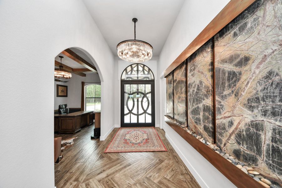 Elegant foyer with a striking circular glass front door, artistic wall feature, and a chandelier, leading to a cozy office space with beamed ceilings.