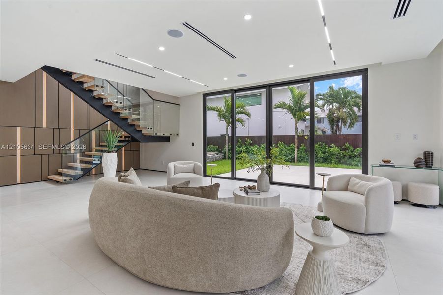 Open-concept interior featuring large-format tile flooring, a custom metal staircase with a glass railing, built-in decorative wall paneling, and floor-to-ceiling sliding glass doors