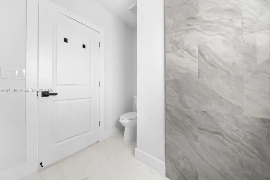 Restroom featuring a wall-mounted door with matte black hardware, large-format tile wall cladding, and polished stone-finish floor tiling Restroom featuring a wall-mounted door with matte black hardware, large-format tile wall cladding, and polished stone-finish floor tiling