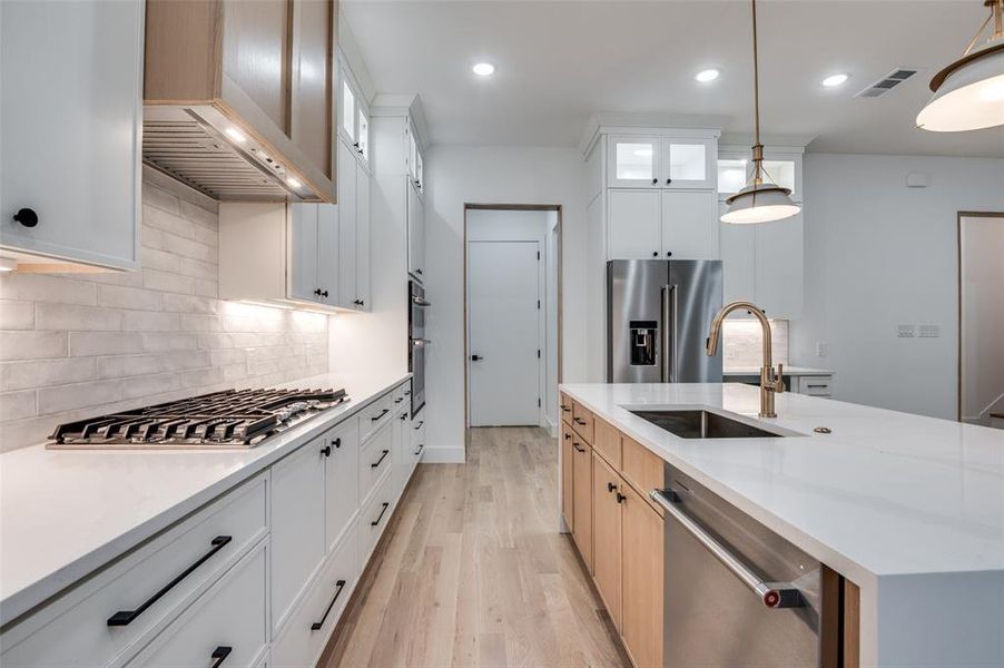 Kitchen featuring light wood finished floors, appliances with stainless steel finishes, a kitchen island with sink, glass insert cabinets, and pendant lighting