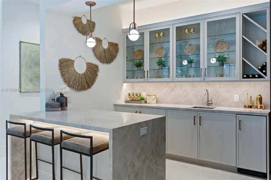 Custom cabinetry with glass-front display doors and wine storage, stone-topped island with waterfall edge, herringbone-patterned tile backsplash, undermount sink, and dual metallic pendant lighting Custom cabinetry with glass-front display doors and wine storage, stone-topped island with waterfall edge, herringbone-patterned tile backsplash, undermount sink, and dual metallic pendant lighting