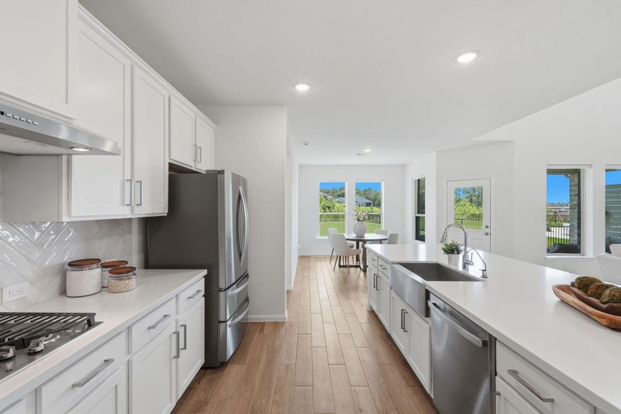 This modern kitchen features sleek white cabinetry, stainless steel appliances, and a spacious island with a farmhouse sink. The open layout flows into a bright dining area with large windows and a view of the backyard, creating an inviting and functional space.