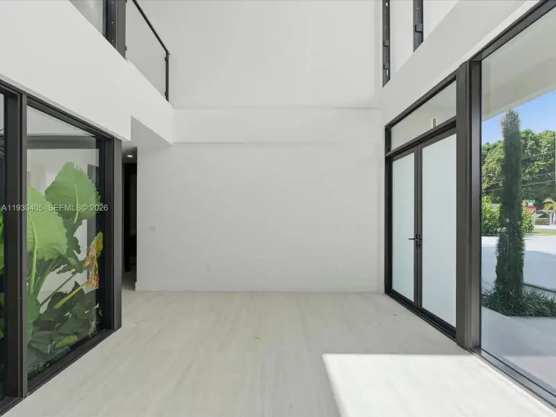 Double-height volume featuring floor-to-ceiling glass windows, full-length stone-finish flooring, metal-framed double glass entry doors, and a glass-paneled mezzanine railing Double-height volume featuring floor-to-ceiling glass windows, full-length stone-finish flooring, metal-framed double glass entry doors, and a glass-paneled mezzanine railing