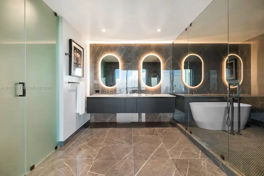 Floating dark wood-finish vanity with dual sinks, backlit oval mirrors, stone-tiled walls and flooring, glass-enclosed walk-in shower, and freestanding soaking tub