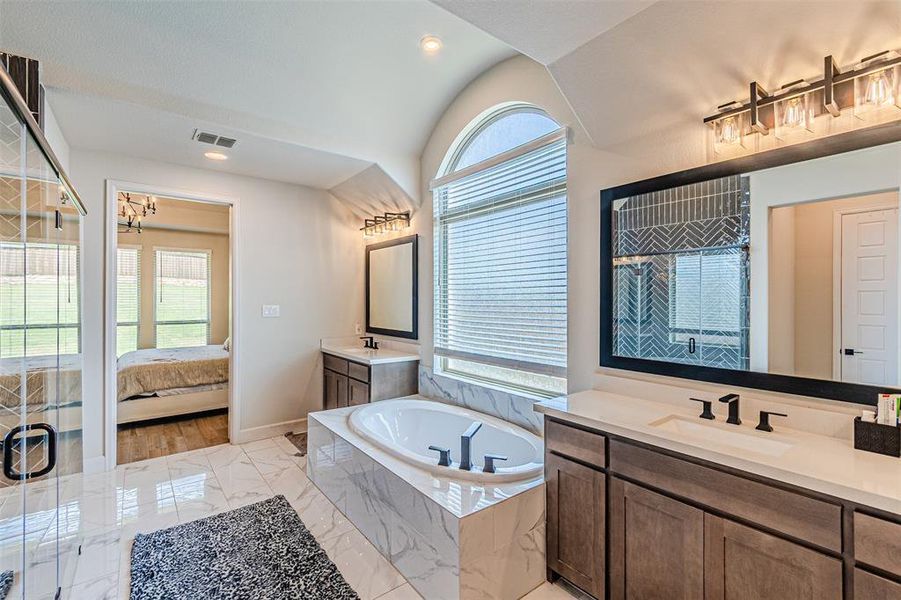 Spacious bathroom featuring a built-in soaking tub with marble-look tile surround, dual vanities with white countertops and dark cabinetry, a large arched window with blinds, and a glass-enclosed shower