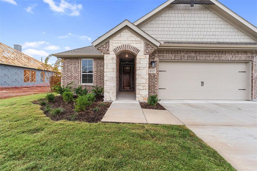 View of front facade featuring brick siding, a shingled roof, concrete driveway, a front lawn, and stone siding View of front facade featuring brick siding, a shingled roof, concrete driveway, a front lawn, and stone siding