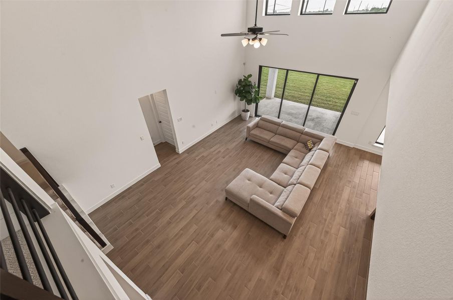 This spacious living room features high ceilings, large windows for natural light, wood flooring, and a modern ceiling fan. A comfortable sectional sofa overlooks a patio and green lawn, creating an inviting, open atmosphere.