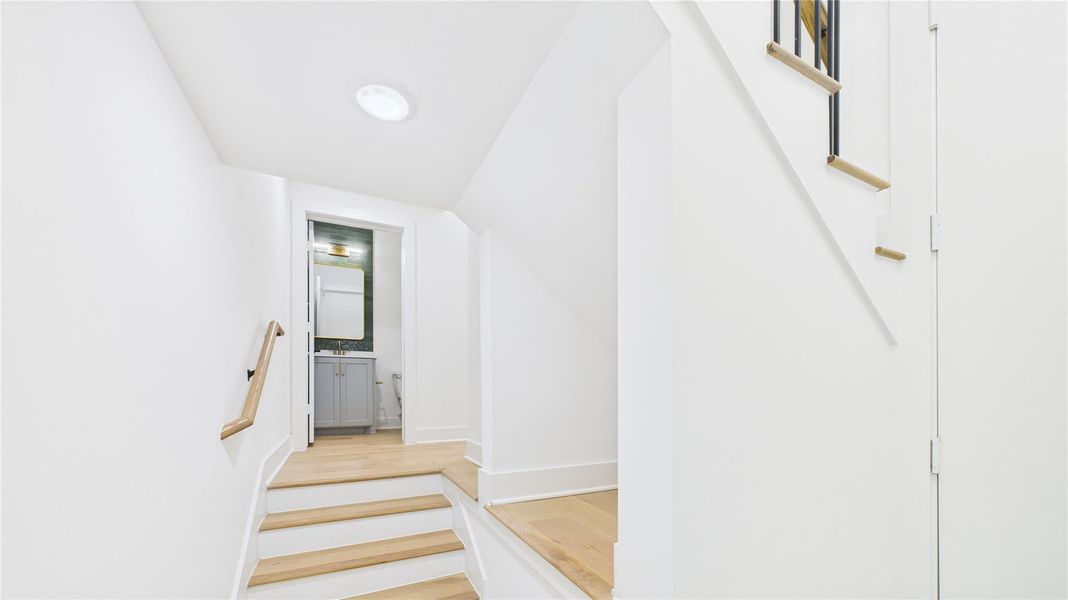 Bright and modern hallway with light wooden steps, white walls, and a sleek handrail, leading to a bathroom with a stylish vanity and mirror.
