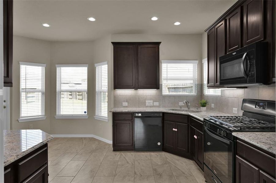 This is a modern kitchen with dark wood cabinets, granite countertops, gas range, and Samsung Fridge is included. It features ample natural light with plenty of windows and is finished with tile flooring.