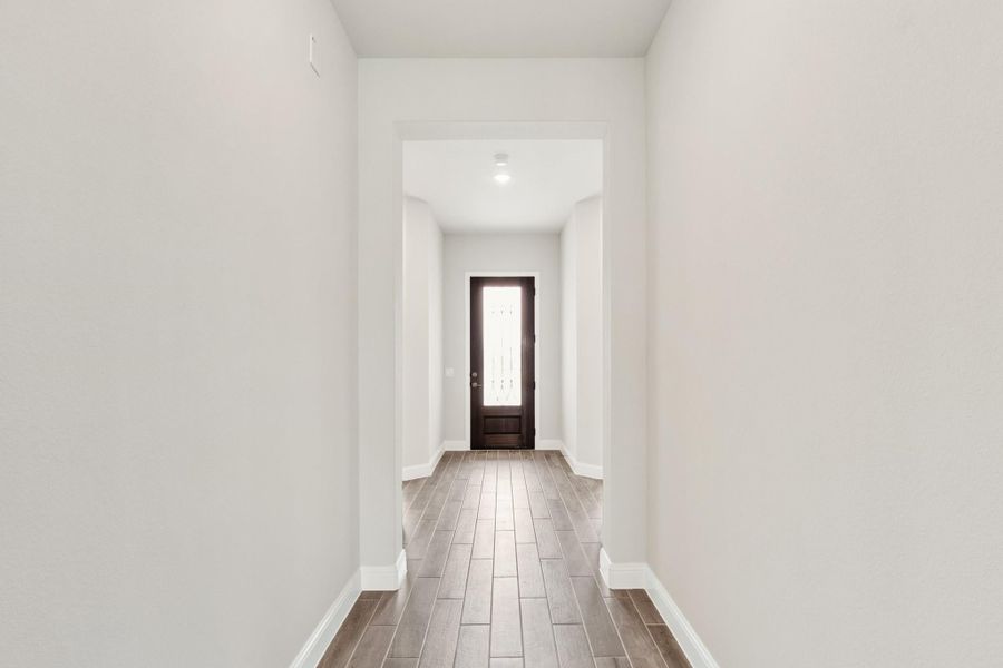 Narrow hallway with wood-look tile floors, white walls, and dark front door with glass panel