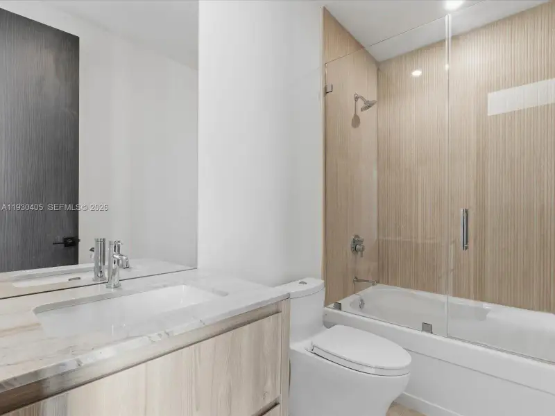 Bathroom featuring a wood-finish vanity with a stone countertop, an integrated rectangular sink, a glass-enclosed shower and tub combination, and vertical-grain wall tile Bathroom featuring a wood-finish vanity with a stone countertop, an integrated rectangular sink, a glass-enclosed shower and tub combination, and vertical-grain wall tile