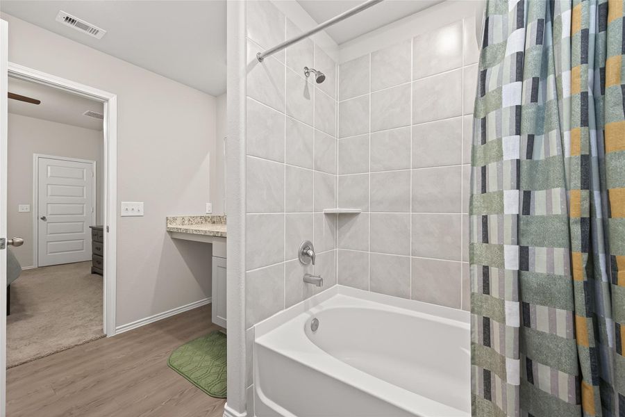 This bathroom features a modern design with a tiled shower and tub combination, accented by a colorful shower curtain. There's a granite countertop with under-sink storage, and the space opens to a carpeted bedroom. The room is bright and has a clean, contemporary feel.
