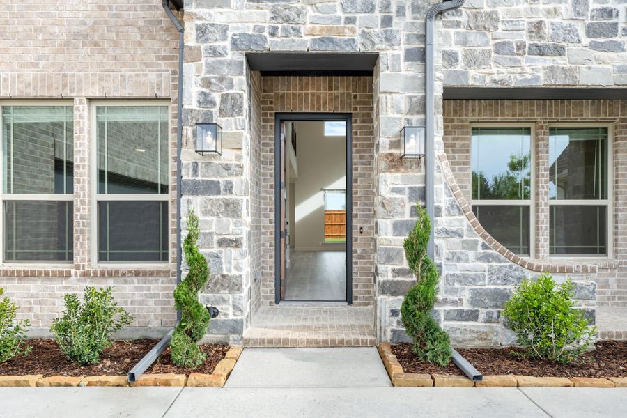 Home entrance with stone and brick exterior, open front door, wall lanterns, and small evergreen shrubs along walkway Home entrance with stone and brick exterior, open front door, wall lanterns, and small evergreen shrubs along walkway