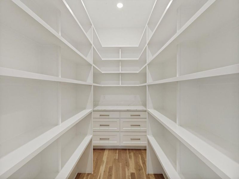 Walk-in pantry featuring extensive white shelving, a built-in cabinet with drawers, a light-toned countertop, and wood-finish flooring Walk-in pantry featuring extensive white shelving, a built-in cabinet with drawers, a light-toned countertop, and wood-finish flooring